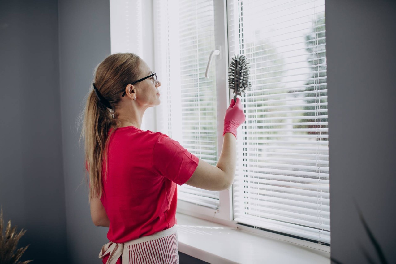 Woman in a red shirt wearing pink rubber gloves doing some blind cleaning with a duster at Delux Drapery & Shade Co. near Ann Arbor, MI